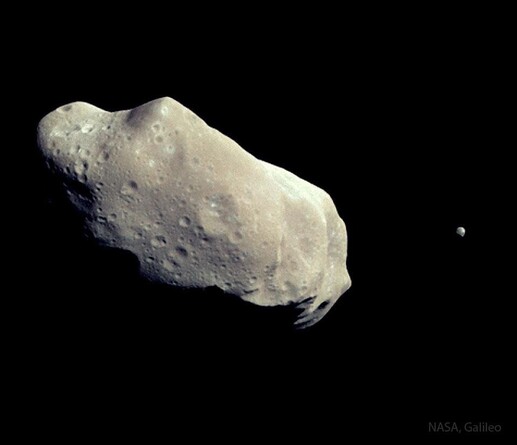 A pair of asteroids are shown with a large, elongated and cratered one on the left and a much smaller one on the far right.