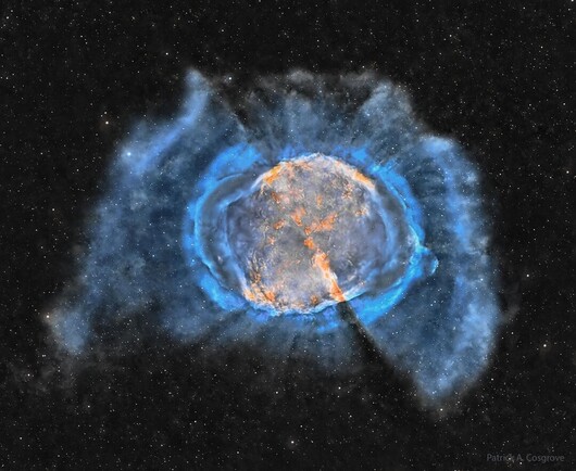 An expansive interstellar gas cloud is shown with an orange interior and outer blue filaments. Many stars are visible in the dark background.